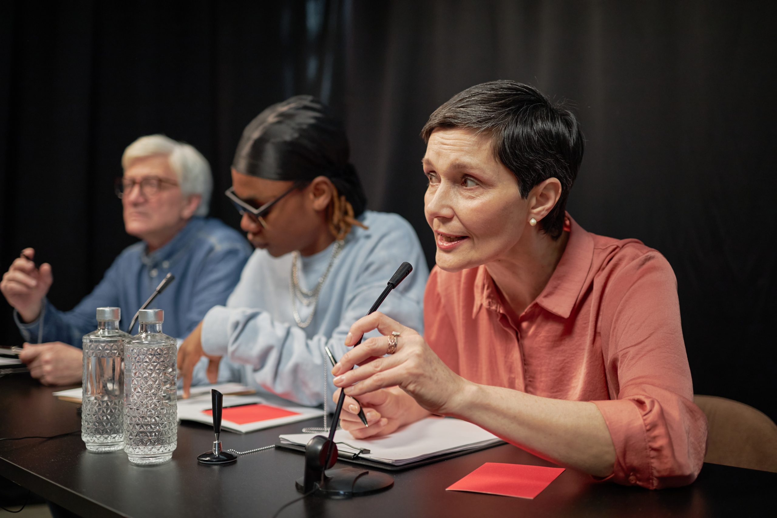 Middle aged Caucasian woman speaking into microphone while sitting at judging table with senior Caucasian man and young Black man during talent competition, all focused on evaluating performances